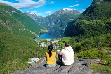 İki kişi Norveç 'te yeşil yemyeşil dağlar ve açık mavi bir gökyüzü ile çevrili pitoresk bir fiyorta tepeden bakan kayalıklarda oturuyor. Geiranger Fjord Norveç