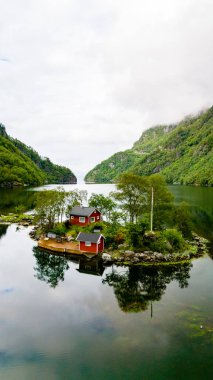 Yeşil ağaçların arasına yuva yapmış iki kırmızı kulübesi olan küçük bir ada, bir fiyortta oturur ve dağlar yükselir. Lovrafjorden, Norveç