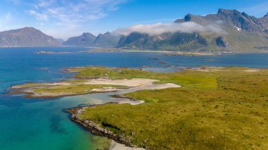 Kumlu kumlu bir sahili olan, temiz mavi su ve uzak dağlarla çevrili küçük, yeşil bir adanın havadan görüntüsü. Kolbedeliden Sahili, Lofoten