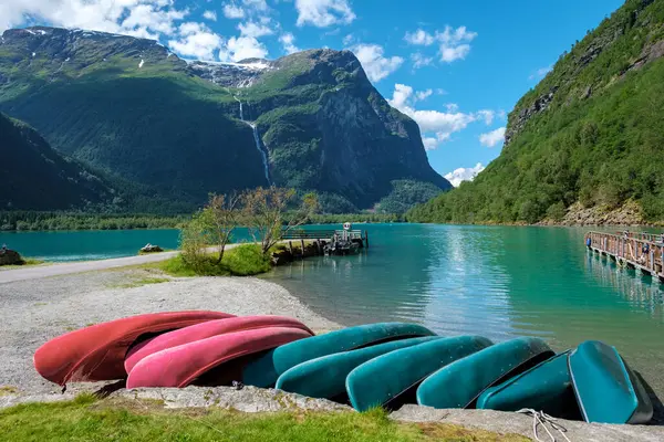 Stock image A row of colorful canoes rests on the shore of a fjord in Norway, surrounded by lush green mountains and a clear blue sky. Lovatnet lake Lodal valley.