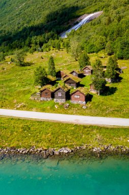 Arka planda çağlayan bir şelale ve turkuaz göle doğru giden dolambaçlı bir yol bulunan yemyeşil bir vadide yuva yapmış geleneksel otlaklı kulübeler. Lovatnet Gölü Lodal Vadisi Norveç