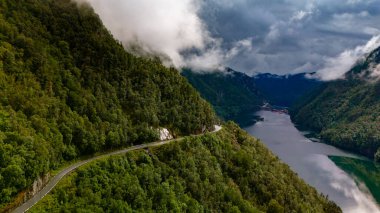 Dönek bir yol, aşağıdaki vadide manzaralı bir göl bulunan yemyeşil bir dağ yamacını kesiyor. Bulutlar gökyüzünde toplanıyor ve manzaraya çarpıcı bir dokunuş katıyor. Lovrafjorden, Norveç