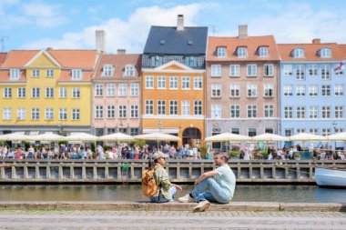 Kopenhag 'da bir kanal boyunca kaldırım kenarında oturan iki kişi gün ışığının ve renkli binaların manzarasının tadını çıkarıyorlar. Kopenhag Danimarka
