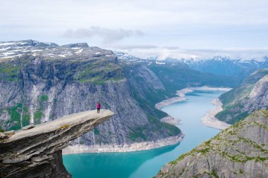 Norveç, Trolltunga 'nın kıyısında yalnız bir yürüyüşçü duruyor. Norveç' in nefes kesici manzarasında sakin bir turkuaz göle bakan dramatik bir uçurum..