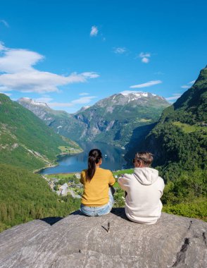 Genç bir çift, yemyeşil ve görkemli dağlarla çevrili, nefes kesici Norveç fiyorduna tepeden bakan kayalık bir çıkıntıda oturuyor. Geiranger Fjord Norveç 'te çeşitli kadın ve erkekler