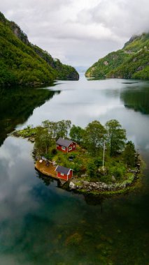 Two red cabins sit nestled on a small, green island in a Norwegian fjord. The water is calm and reflects the sky and the surrounding mountains. Lovrafjorden, Norway