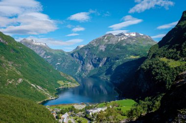Sakin mavi bir su kütlesini çevreleyen yemyeşil dağlarla Norveç fiyordunun manzarası. Gökyüzü açık ve mavi, tüylü beyaz bulutlarla. Geiranger Fjord Norveç