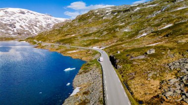 Norveç 'te manzaralı bir dağ geçidini takip eden dolambaçlı bir yol, ışıl ışıl mavi bir göl ve karla kaplı tepeleri var. Yol boyunca bir karavan sürüyor, Strynefjellsvegen, Geiranger, Norveç