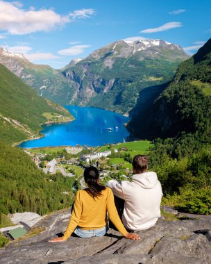 Bir çift, yemyeşil ve görkemli dağlarla çevrili, göz kamaştırıcı bir Norveç fiyorduna bakan kayalık bir çıkıntıda oturuyor. Geiranger Fjord Norveç 'te çeşitli kadın ve erkekler var.
