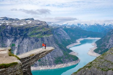 İki yürüyüşçü, Norveç, Trolltunga 'daki dramatik fiyort ve dağ manzarasına bakan bir uçurumun kenarında duruyor.