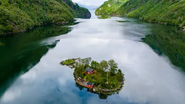 stock image a small island nestled within a picturesque Norwegian fjord. Lush greenery adorns the island, with a cozy red cabin adding a touch of warmth to the serene landscape. Lovrafjorden, Norway