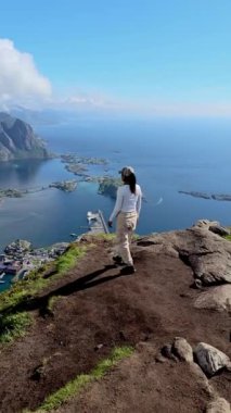 Göl kenarındaki dağlarla çevrili bir kasabayı görüntüler. Bir kadın tepeden gelen suların güzelliğine hayran. Doğanın en güzel manzarası burada, Reinebringen, Lofoten Norveç