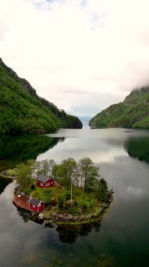Havadan bakıldığında, yeşil ağaçlarla sarılmış küçük bir adası olan sakin bir göl görülüyor. Adadaki resim gibi kırmızı bir ev huzurlu ortamı güçlendirir. Lovrafjorden, Norveç