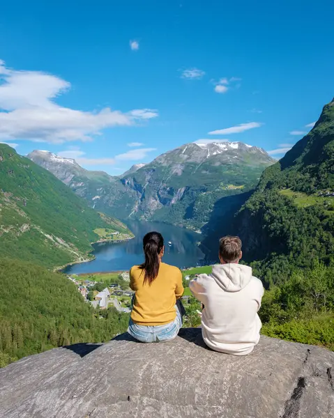 stock image A couple sits on a rock, gazing at the breathtaking panorama of a Norwegian fjord, surrounded by lush green mountains under a clear blue sky. diverse men and women at Geiranger Fjord Norway