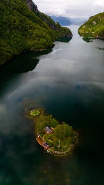 A small, forested island with a couple of red cabins sits in the middle of a deep, dark fjord, framed by towering green cliffs. Lovrafjorden, Norway