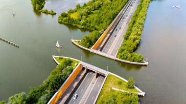 Hollanda 'daki Aquaduct Veluwemeer' in hava görüntüsü, su kanalının altından geçen bir otoyol gösteriyor. Otoyol yemyeşil ağaçlar ve çalılarla çevrili..