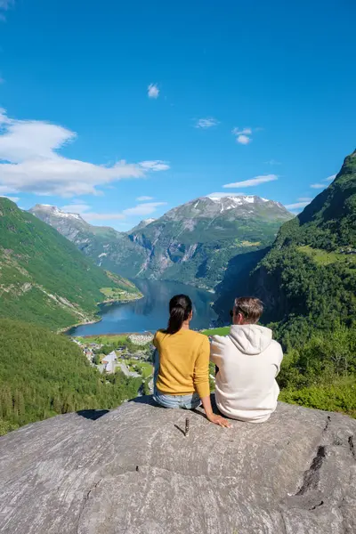 stock image Two people sit on a rock overlooking a picturesque Norwegian fjord, surrounded by lush green mountains and a clear blue sky. a diverse couple of men and women at a viewpoint Geiranger Fjord Norway