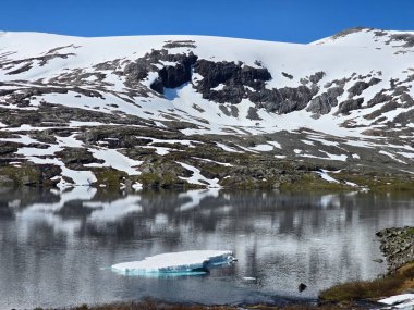 Huzurlu bir manzara, pırıl pırıl mavi gökyüzünün altındaki görkemli karla kaplı tepeleri yansıtan kristal berraklığında bir göl sergiliyor..