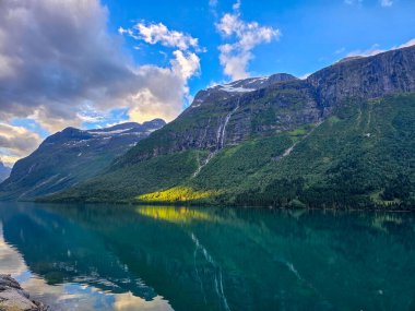 Engebeli dağlarla çevrili sakin bir fiyordun sakin sularında altın ışık dans eder ve dramatik bir gökyüzü altında yemyeşil. Lovatnet, Norveç