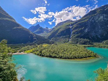 Bereketli yeşillik, Norveç 'in çarpıcı manzarasında yükselen dağlar ve parlak bulutlarla çerçevelenmiş bir fiyortun parıldayan turkuaz sularını kucaklıyor. Lovatnet, Norveç 