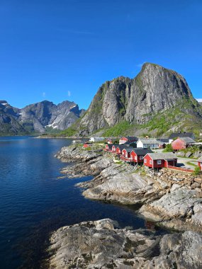 Kayalık kıyı boyunca yuva yapmış canlı kırmızı kulübeler, berrak bir gökyüzünün altında görkemli dağlarla çevrili sakin sulara karşı yansıyor. Lofoten Norveç 