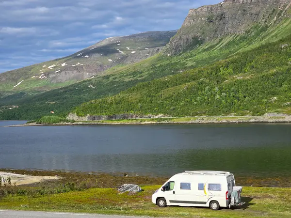 stock image A cozy camper van rests beside a tranquil lake, surrounded by lush greenery and towering mountains under a bright sky.