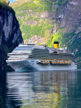 Geiranger Norveç 'e 10 Temmuz 2024' te şafak sökerken Norveç 'in fiyortlarının sakin sularında yol alan çarpıcı bir yolcu gemisi yükselen kayalıklar ve canlı yeşilliklerle çevrilidir..