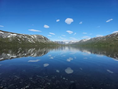 Kristal berrak göl görkemli dağları ve kabarık bulutları yansıtıyor, güneşli bir günde Norveç 'in huzurlu güzelliğini yakalıyor..