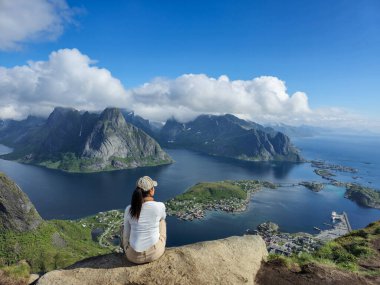 Bir insan, Norveç 'in Lofoten kentindeki dağların nefes kesici manzarasına bakarak kayalık bir çıkıntıda oturur. Şafağın yumuşak ışığıyla aydınlanır. Reinebringen Lofoten Norveç 
