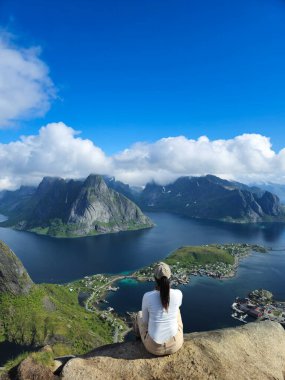 Tek bir figür, Norveç 'in çarpıcı Lofoten Adaları' ndaki yemyeşil dağların ve sakin suların nefes kesici panoramik manzarasına hayran kalarak kayalık bir çıkıntıda rahatlar..