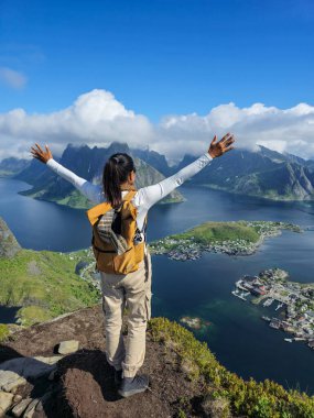 Bir yürüyüşçü bir uçurumun tepesinde duruyor, kolları ardına kadar açık, temiz mavi gökyüzünün altındaki fiyortların ve adaların nefes kesici manzarasının tadını çıkarıyor. Reinebringen Lofoten Norveç 