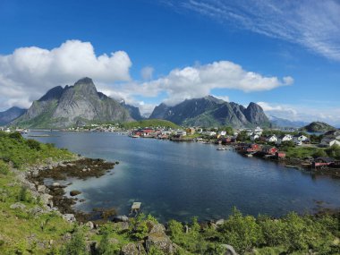Parlayan sular kıyı şeridi boyunca yükselen dağlar ve berrak mavi gökyüzü tarafından çerçevelenmiş canlı evleri yansıtıyor. Reine, Lofoten Norveç 