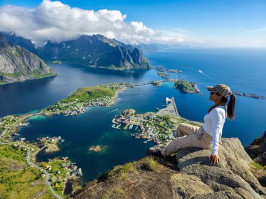Bir maceraperest, Lofoten Adaları 'nın panoramik güzelliğinde, sakin suları ve engebeli dağları aydınlatan kayalık bir çıkıntıda oturur. Reinebringen Lofoten Norveç 
