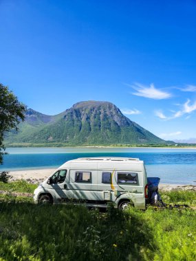 Rahat bir karavan, görkemli dağlar ve yemyeşil yeşilliklerle çevrili engin gökyüzünün altında parıldayan mavi sularda dinleniyor. Lofoten Norveç 