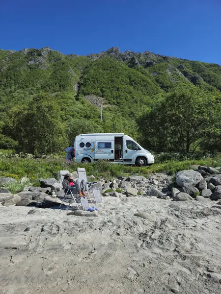 stock image A camper van rests against a backdrop of lush green mountains, while a person relaxes in a beach chair, enjoying the tranquility of nature. Lofoten Norway 