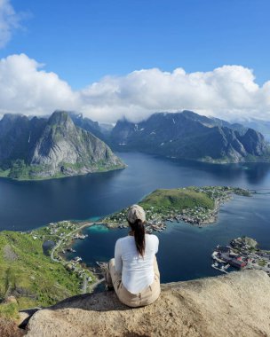 Bir gezgin kayalık bir çıkıntıda oturur, göz kamaştırıcı mavi sulara ve parlak bir gökyüzünün altındaki Lofoten Adaları 'nın engebeli dağlarına bakar. Reinebringen Lofoten Norveç 