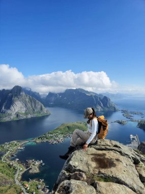Bir gezgin engebeli bir uçurumun tepesinde oturur, aşağıdaki muhteşem fiyorda bakar, görkemli tepeler ve berrak gökyüzü ile çevrilidir. Reinebringen Lofoten Norveç 