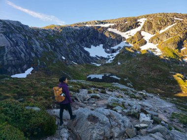 Yalnız bir yürüyüşçü, yüksek dağlar ve kar parçalarıyla çevrili kayalık arazide güneş ışığında güneşlenerek dolaşır. Lysefjord Kjeragbolten Kjerag Norveç