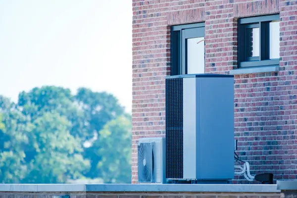 stock image A sleek air source heat pump is mounted on the exterior of a contemporary brick structure, surrounded by lush greenery under a clear sky. warmte pomp translation air source heat pump