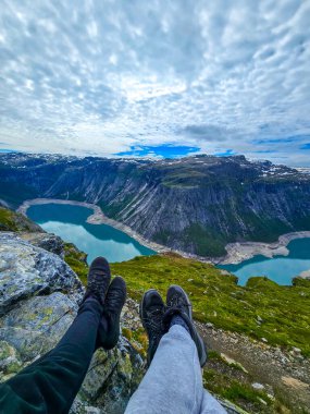 Norveç 'te, bulutlarla boyanmış bir gökyüzü altında yemyeşil ve dramatik uçurumlarla çevrili nefes kesici bir fiyordun üzerinde iki çift ayak sallanıyor.