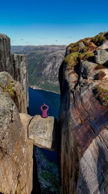 Yalnız bir maceracı Kjeragbolten uçurumunun tepesine uzanır, nefes kesici fiyort manzaraları ve parlak mavi gökyüzünün altındaki engebeli dağlarla çevrili. Kjerag Kjeragbolten Lysefjord Norveç