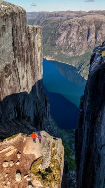 İki yürüyüşçü dramatik bir uçurumun kenarında dikilip aşağıdaki derin mavi fiyorda bakıyor ve parlak gökyüzünün altında etraflarındaki kaya oluşumlarını izliyorlar. Kjerag Kjeragbolten Lysefjord Norveç