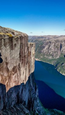 Parlak bir gökyüzünün altında yemyeşil dağlarla çevrili derin mavi fiyort üzerinde nefes kesen uçurumlar dramatik bir şekilde yükselir. Kjerag Kjeragbolten Lysefjord Norveç