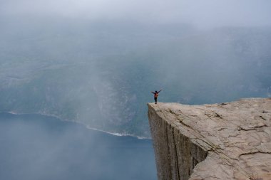 Solo bir yürüyüşçü zaferle Preikestolen uçurumunda poz verir, puslu gri gökyüzü ve aşağıdaki çarpıcı fiyortlarla çevrilidir. Önceki Norveç