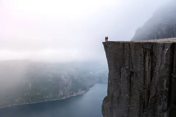 stock image Amidst misty surroundings, two adventurers stand at the edge of Preikestolen Cliff, gazing at the stunning Lysefjord below, embracing nature's grandeur and tranquility. Preikestolen Norway