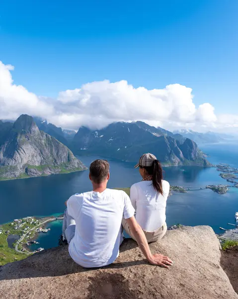 stock image A couple sits on a rocky ledge, gazing out over the serene waters and dramatic mountains of the Lofoten Islands under a bright, blue sky. Reinebringen hike Lofoten Norway