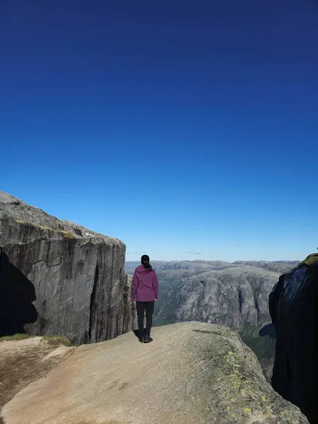 Stock image A solitary figure stands atop Lysefjord Kjeragbolten Norway, gazing out over the breathtaking fjords and rugged mountains of Norway under a vibrant blue sky.