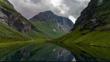 Yükselen dağlar, üzerinde süzülen kara bulutlar ile sakin bir fiyort çerçeveler, durgun su üzerinde dramatik bir yansıma bırakarak.