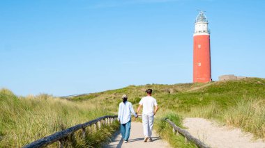 Texel Hollanda, kumlu sahilde yükselen Majestic deniz feneri güneşli bir günde deniz fenerine yürüyen bir çift adam ve kadın.
