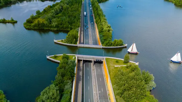 Stock image A road stretches across a canal, creating a unique bridge over water. Lush green foliage borders the waterway and a small sailboat drifts peacefully nearby. Veluwemeer Aquaduct in the Netherlands